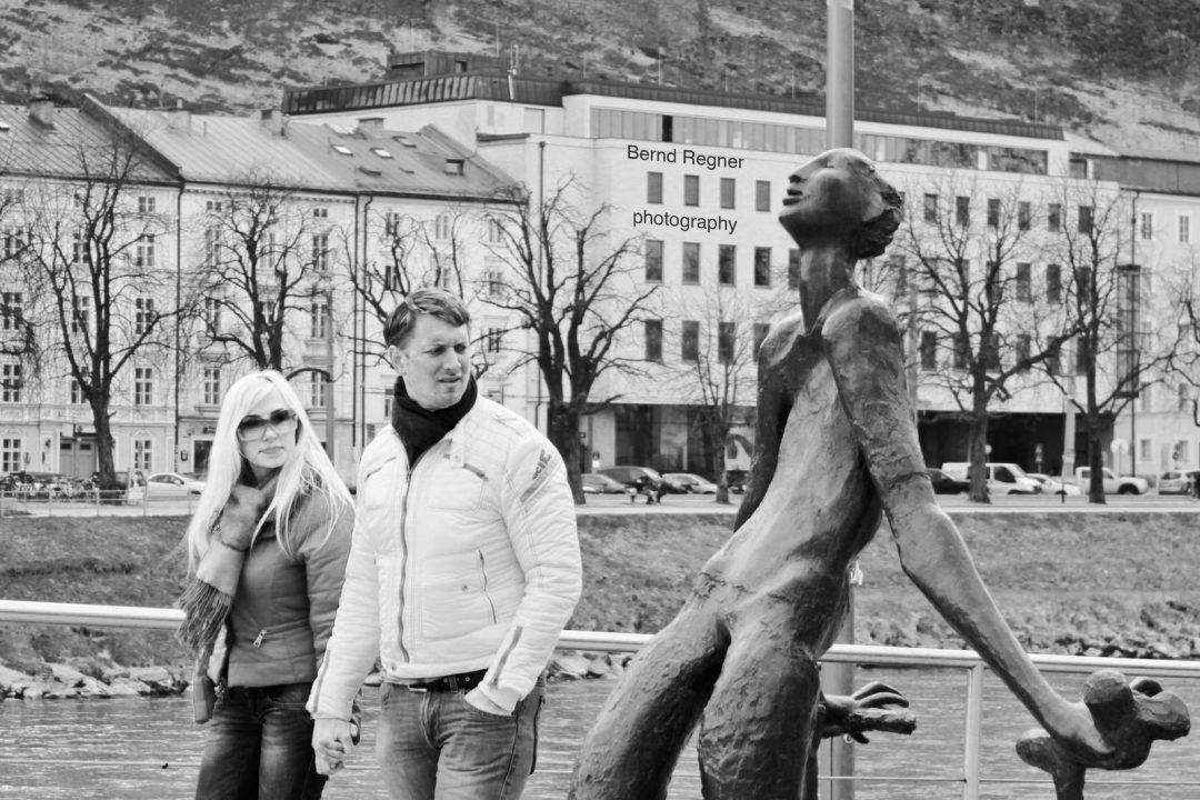 black and white photo of a statue by the river Salzach, being looked at in a weird way by a couple